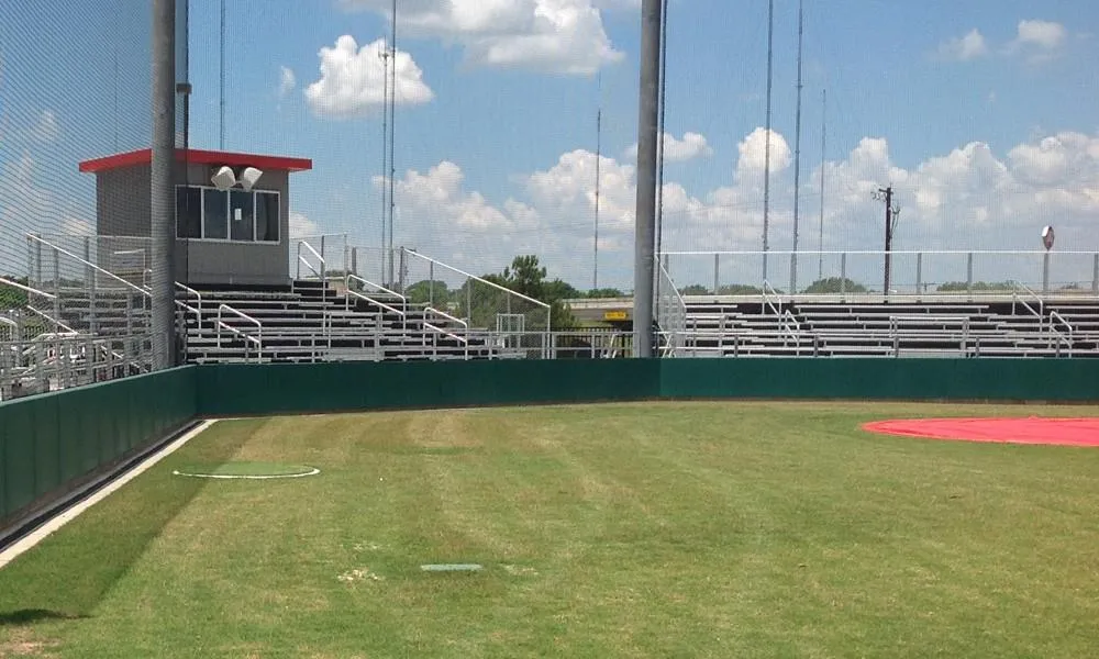 A baseball field with a well-maintained grassy outfield, surrounded by tall netting. In the background, there are empty bleachers and a small building with a red roof under a blue sky with a few clouds.