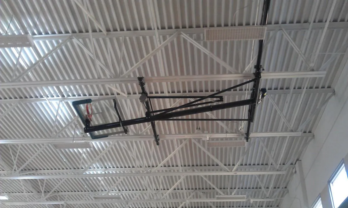 A metal basketball hoop is mounted on the ceiling of a large indoor gymnasium. The ceiling features white, corrugated metal panels and exposed steel beams. The hoop is retracted horizontally.