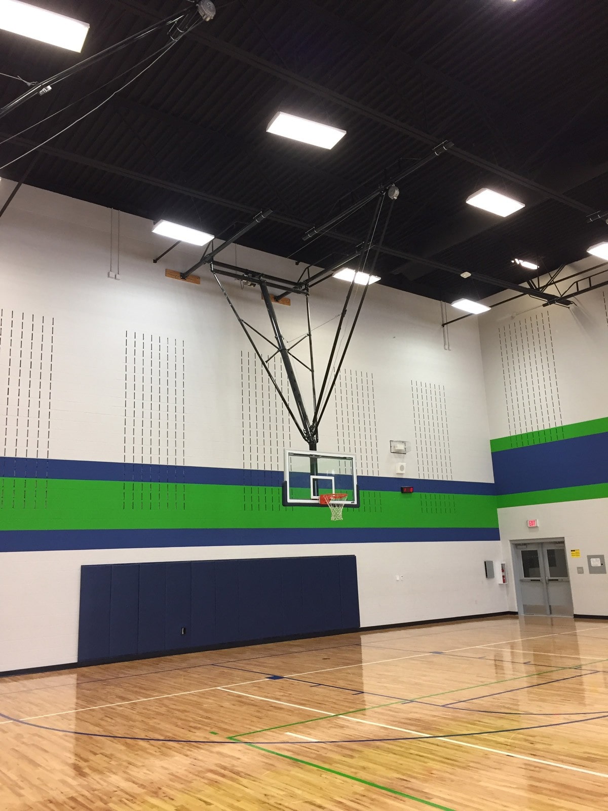 Indoor basketball court with a wooden floor, a basketball hoop and backboard mounted on the wall. The walls have green and blue stripes, and there are acoustic panels and ceiling lights visible.