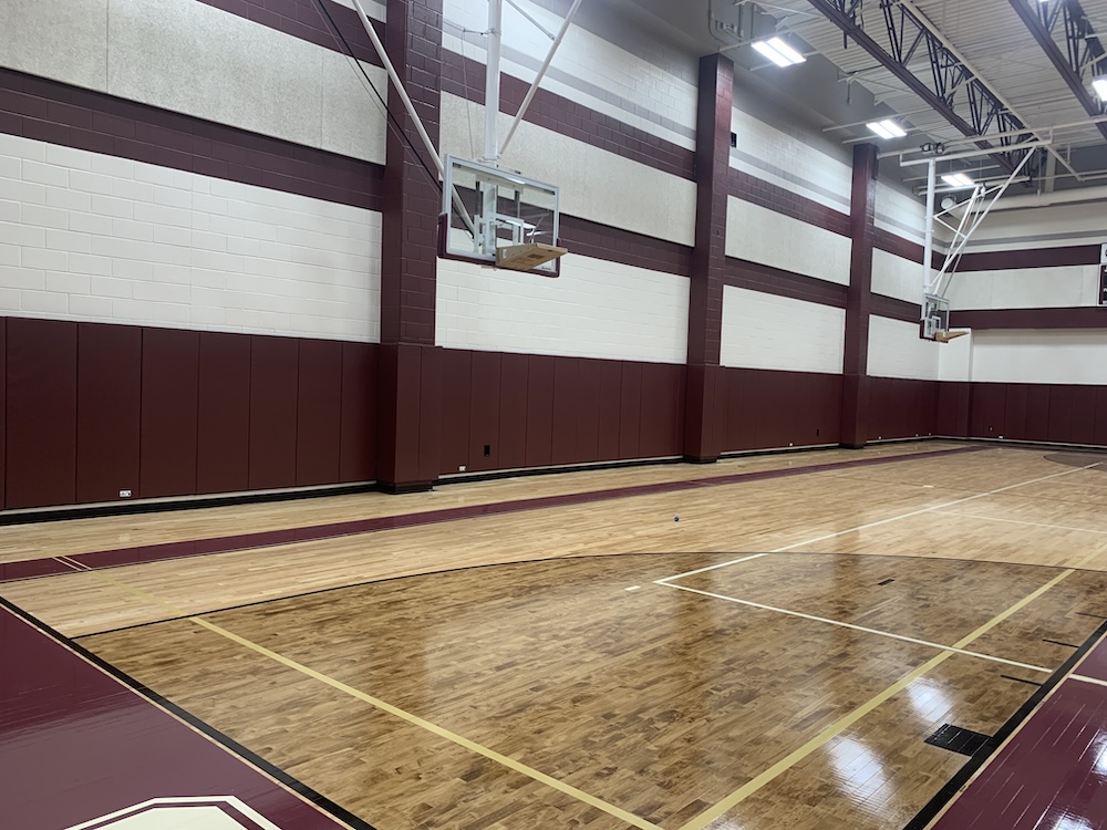 Wide-angle view of an empty indoor basketball court with polished wooden floors and maroon padded walls. Ceiling lights illuminate the court, and basketball hoops are visible on the left. The space is well-maintained and ready for use.