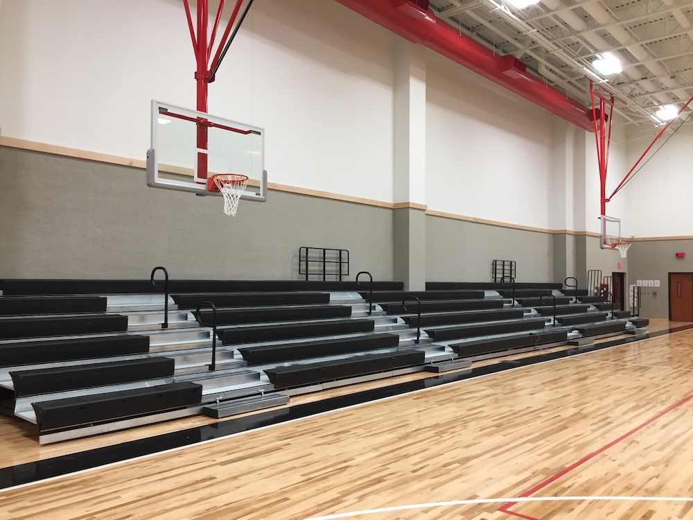 Empty indoor basketball court with wooden flooring, featuring red and black bleachers along the wall. The ceiling has red support beams and lighting, and there are basketball hoops without nets on either side.