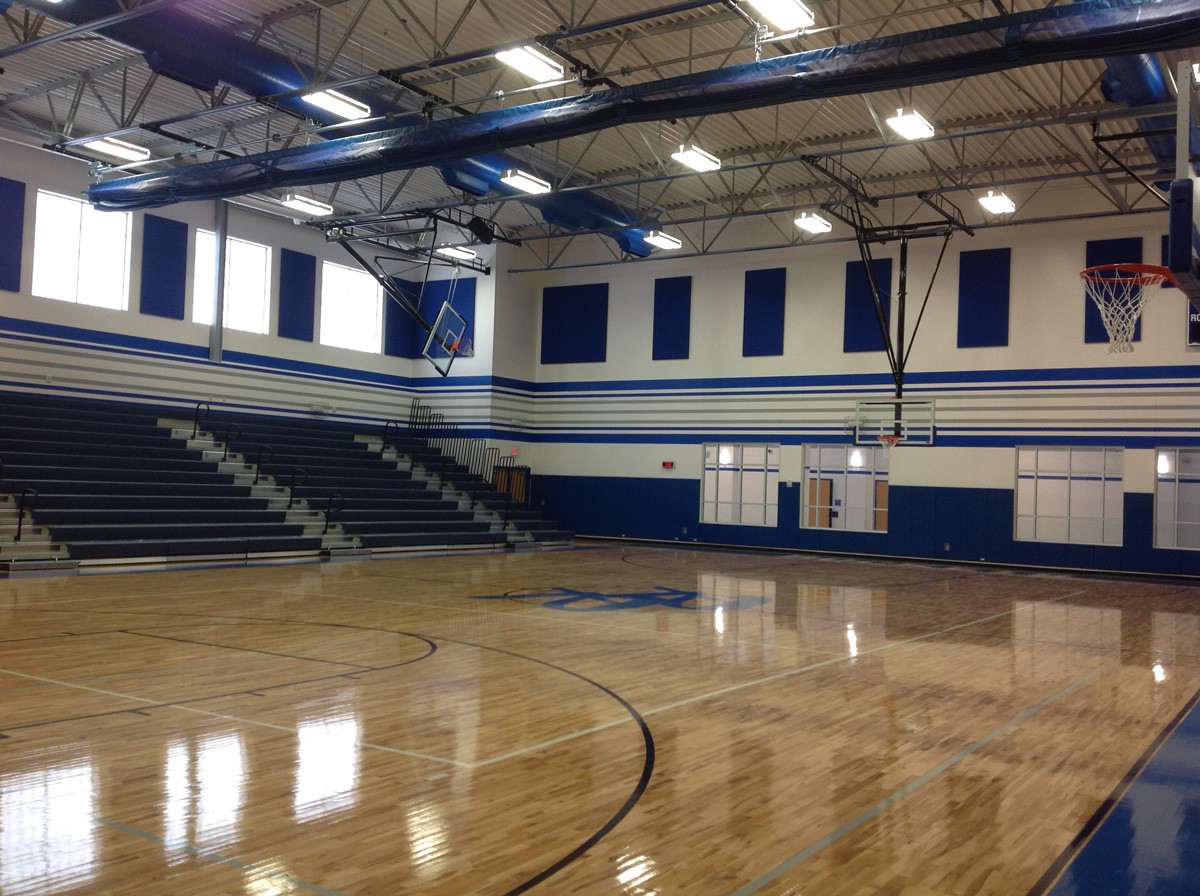 An empty gymnasium with polished wooden floors. Multiple basketball hoops are mounted on the walls, and bleachers are on one side. The walls and ceiling feature blue and white accents, with natural light streaming in through large windows.