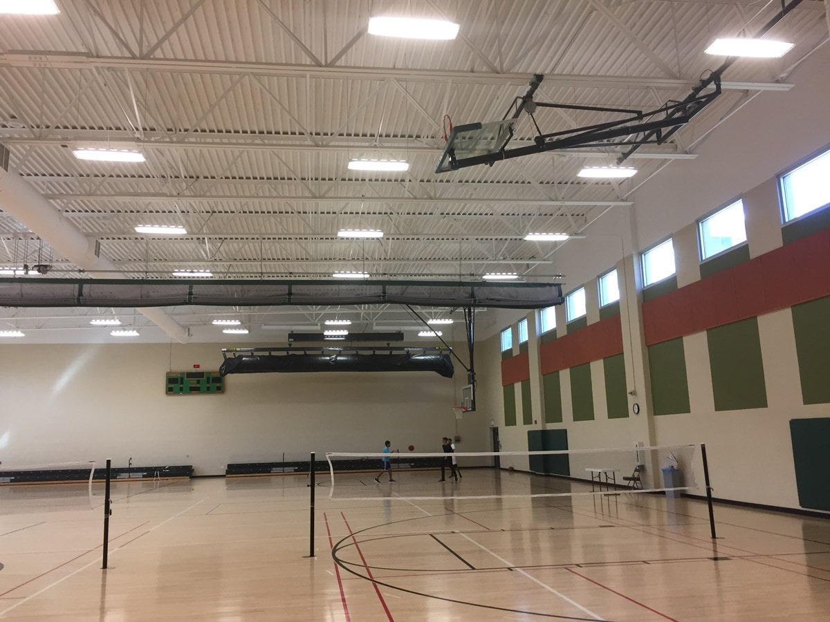 A spacious indoor gym with a polished wooden floor featuring two badminton courts with nets set up. The ceiling is high with bright lighting, and a few people are playing in the background. The walls have colorful panels near the ceiling.