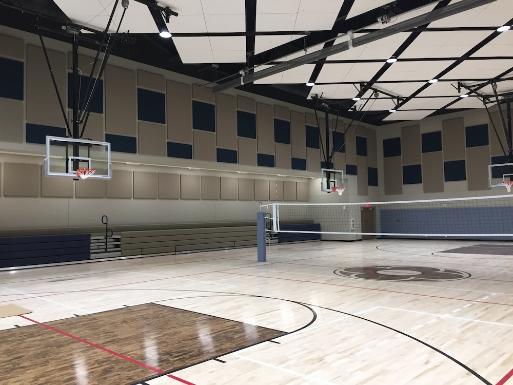 A spacious indoor gymnasium featuring a polished wooden floor with basketball court markings. Two basketball hoops are mounted on opposite ends. A volleyball net is set up in the center, and bleachers line one wall.