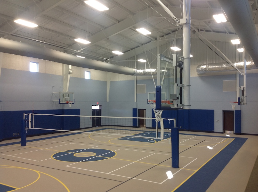 A spacious indoor gymnasium features a basketball court with hoops on either end and a volleyball net set up at the center. The ceiling is high with visible ventilation ducts, and natural light enters through small windows.