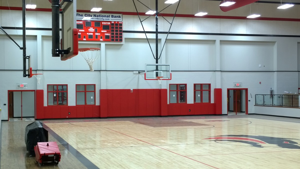 A basketball court inside a gymnasium with red and white walls. A scoreboard displays teams and scores. Red padding lines the walls beneath the hoops. A bench is visible on the side of the court. The ceiling features fluorescent lights.