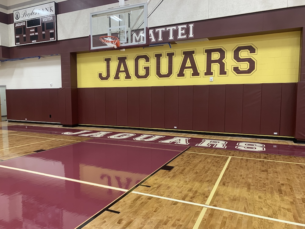 A gymnasium with a polished wooden floor and maroon walls. The wall features bold lettering: "MATTEI JAGUARS" in maroon and yellow. A basketball hoop is mounted on the wall, and a scoreboard is visible in the corner.