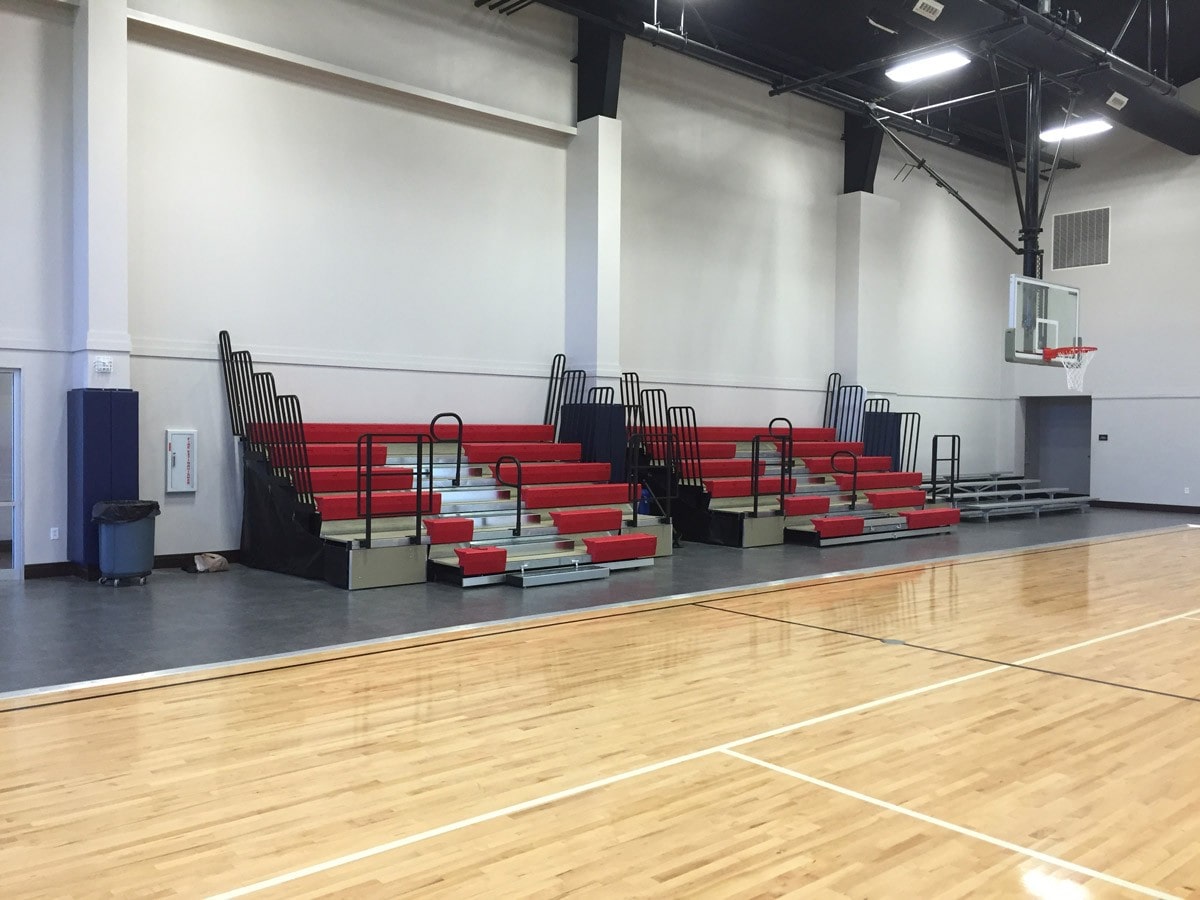 Indoor basketball court with a light wooden floor. On the left side, there are red and black retractable bleachers. A basketball hoop is visible on the right. The space is well-lit with a high ceiling.