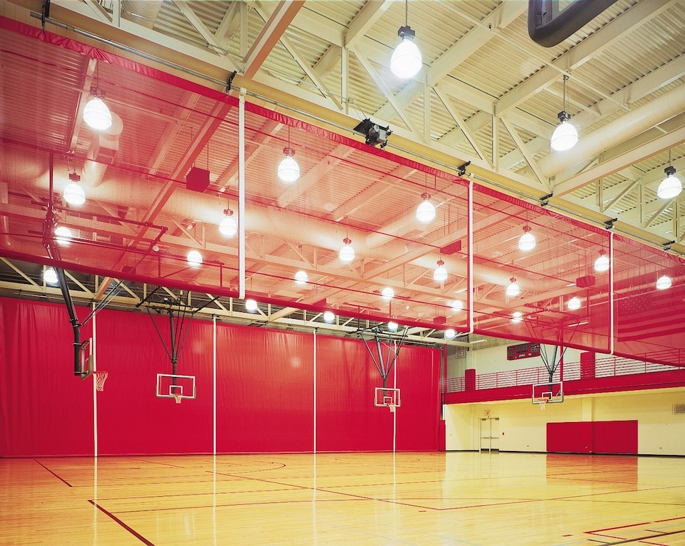 An indoor gymnasium with hardwood floors, featuring multiple basketball courts. Red curtains are drawn across the space, dividing the courts. The ceiling is high with exposed beams and bright overhead lights. Red walls surround the courts.