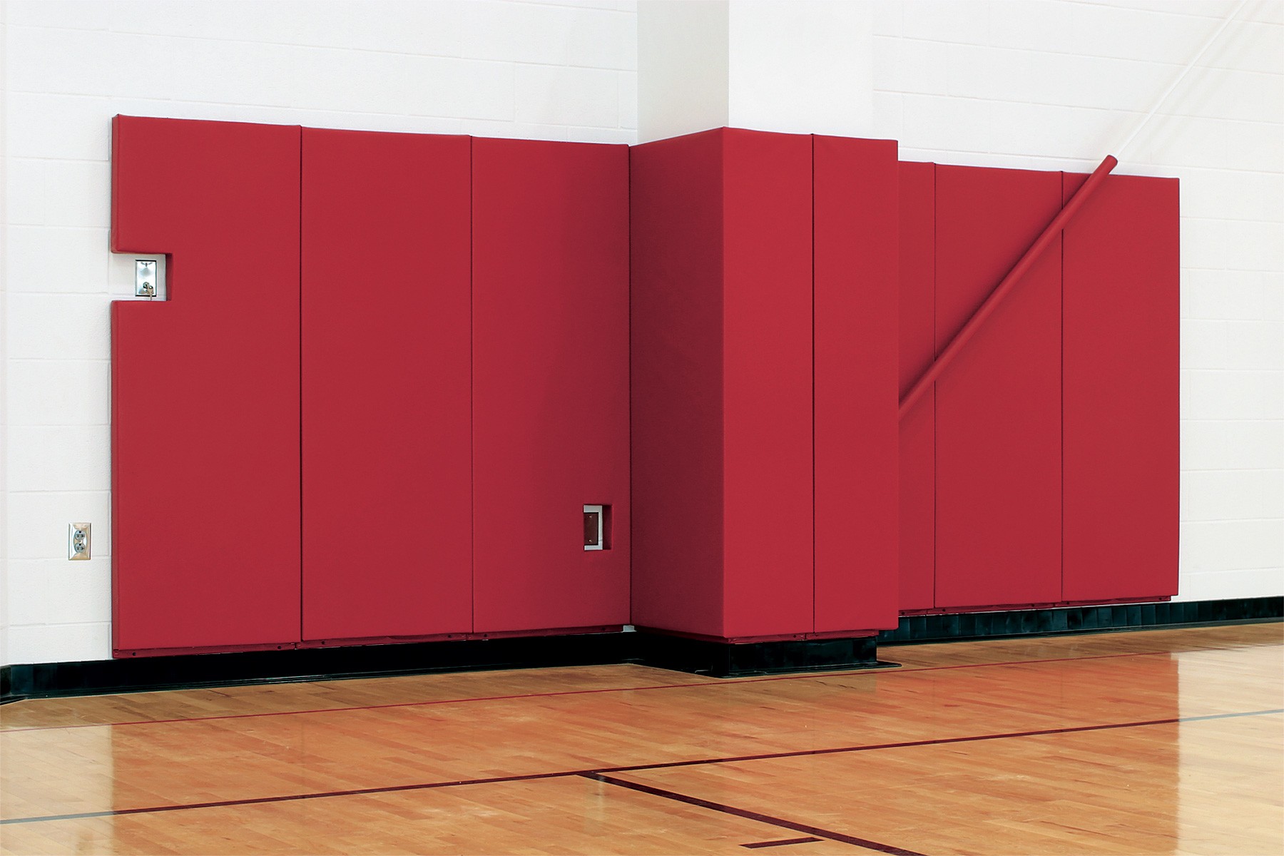 Red wall padding installed on a white gymnasium wall, extending across several panels with a protruding section. The floor is wooden with visible sports court markings.