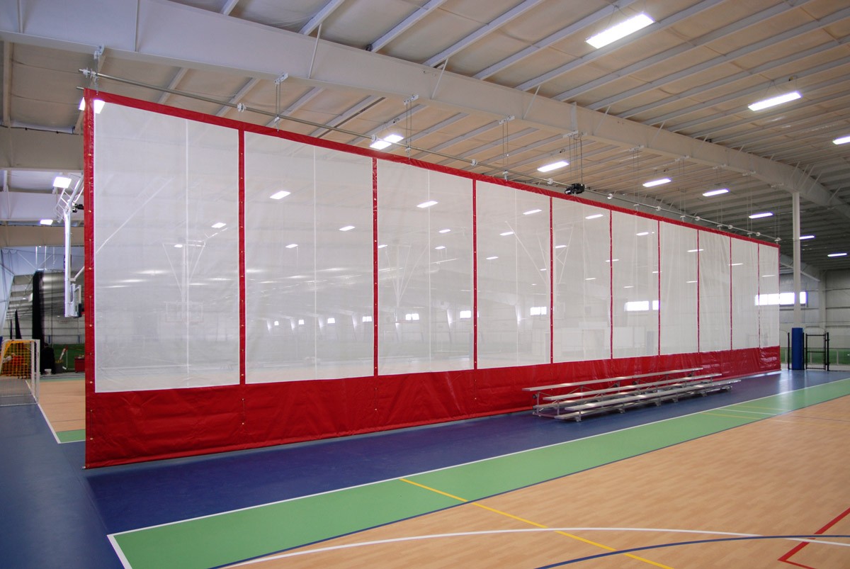 Indoor sports court with a red and white divider curtain. The floor is marked with lines for different sports, including basketball and volleyball. Bleachers are positioned to the side. The ceiling is high with bright overhead lights.