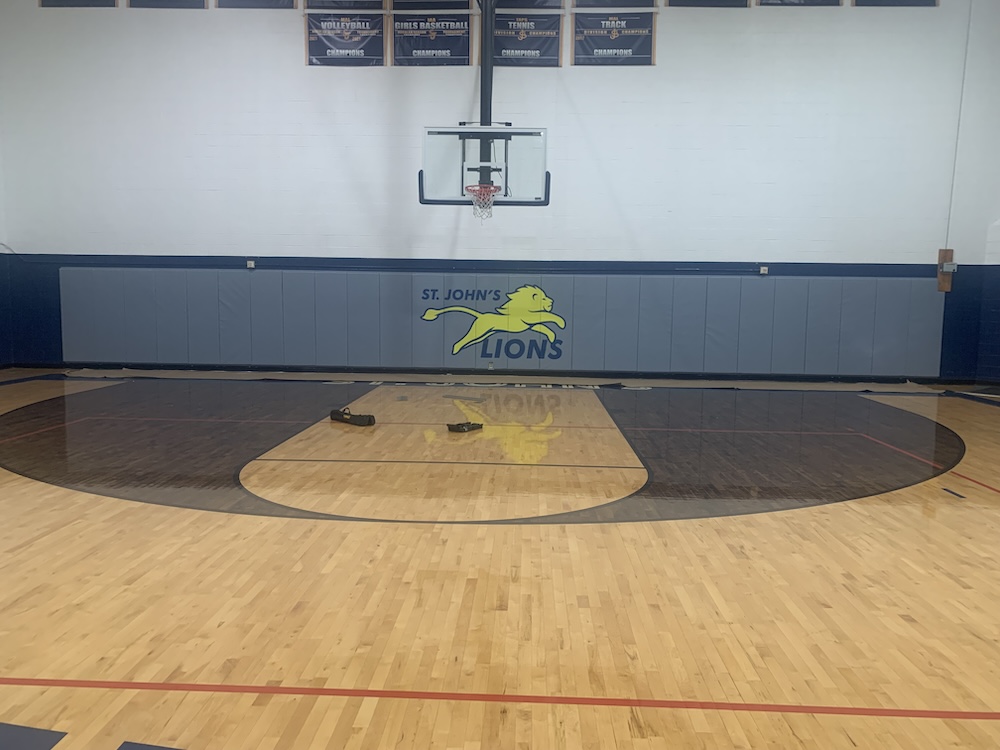 A gymnasium with a basketball hoop and a polished wooden floor. The wall features the St. John's Lions logo, depicting a yellow lion with blue text. Banners hang above the court, and there are pieces of equipment on the ground.