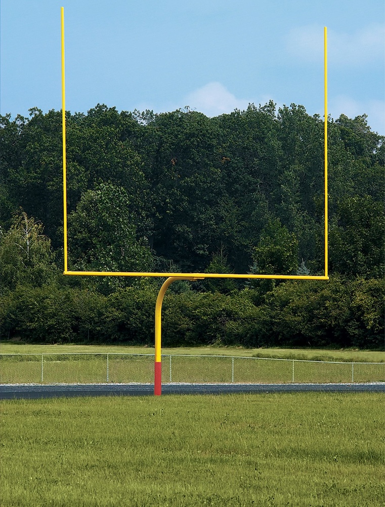 A yellow football goalpost stands in a grassy field with a background of dense green trees under a blue sky.